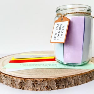 May include: A glass jar filled with coloured paper notes, tied with twine and a tag that reads "Read Me When You Can't Sleep." The jar sits on a wooden slice with additional notes. The notes are various colours, including purple, blue, yellow, red, and green.