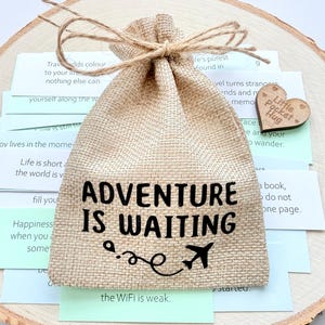 Può includere: Un sacchetto regalo in juta con la scritta "ADVENTURE IS WAITING" e un aereo. Il sacchetto è legato con spago e poggia su una fetta di legno, circondato da piccole carte e un cuore di legno con "Little Pocket Hug".