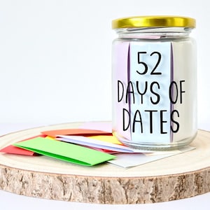 May include: A glass jar with a gold lid, labelled "52 Days of Dates" in black text, rests on a wooden slice. Several colourful folded paper notes are scattered around the jar. The jar is filled with white paper.