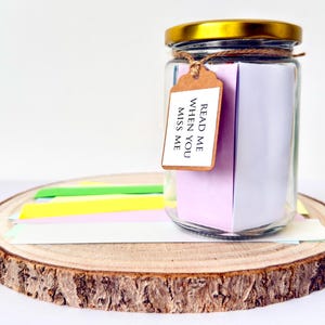 May include: A glass jar with a gold lid and a tag saying "Read Me When You Miss Me." The jar is filled with colourful paper notes and sits on a wooden slice with colourful paper strips.