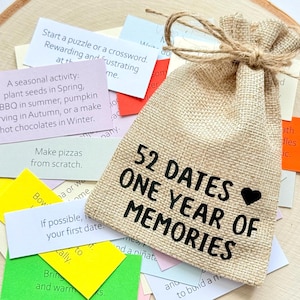 May include: A tan burlap drawstring pouch with the black text 52 DATES ONE YEAR OF MEMORIES and a small heart icon. The pouch is surrounded by a collection of small, rectangular paper slips in various colors, each featuring printed activity prompts.