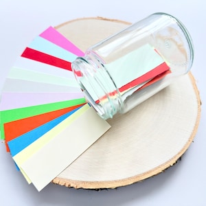 Può includere: Un barattolo di vetro trasparente rovesciato su una fetta di legno, con strisce di carta colorate. Le strisce sono di vari colori, tra cui rosa, rosso, verde, arancione, blu e giallo. Il barattolo contiene strisce di carta simili.