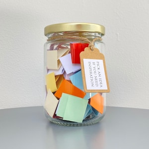 Puede incluir: Un frasco de vidrio lleno de cuadrados de papel de colores, cada uno con una idea escrita en él. El frasco tiene una tapa dorada y una etiqueta marrón que dice "Elige una idea si necesitas inspiración".