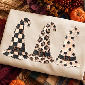 Può includere: Una felpa color crema con una stampa grafica di tre cappelli da strega disposti in fila. Il primo cappello presenta un motivo a scacchi nero e crema, il secondo una stampa leopardata e il terzo pois neri su una base color crema.