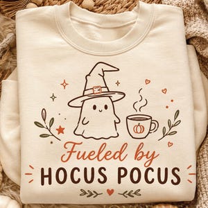 May include: A cream-colored crewneck sweatshirt featuring a graphic of a ghost wearing a witch hat next to a steaming mug with a pumpkin icon. The text reads "Fueled by HOCUS POCUS" in orange and dark brown, surrounded by small hearts, stars, and leaves.