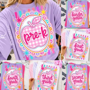 Könnte beinhalten: Ein hellviolettes T-Shirt mit einer zentralen Grafik eines rosa-weiß karierten Apfels in einem gewellten Rahmen mit bunten Sternen und Streifen. Der Apfel enthält den Text 'pre-k' in Schreibschrift, umgeben von weiteren T-Shirts.