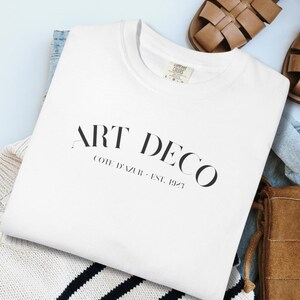 Op de afbeelding: Een wit sweatshirt met ronde hals en de zwarte tekst ART DECO COTE D'AZUR EST. 1923 op de voorzijde. Het sweatshirt ligt op een wit met donkerblauw gestreepte trui, bruine leren sandalen en een bruine handtas van gevlochten leer.