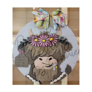 以下が含まれることがあります： ピンクの花の冠をかぶった茶色のハイランド牛の顔が描かれた丸い木製の看板。看板には、茶色のタグに「Live Like Someone Left the Gate Open」という文字が書かれています。