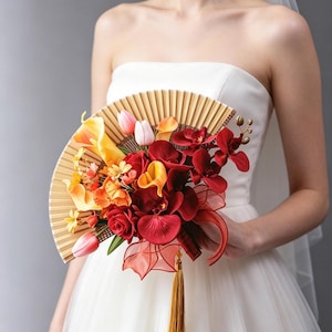 Puede incluir: Un abanico plegable decorado con un arreglo de flores artificiales que incluye rosas rojas, orquídeas rojo oscuro, calas amarillas y naranjas, y tulipanes rosa claro. El abanico presenta una estructura acanalada marrón claro y una borla dorada larga.