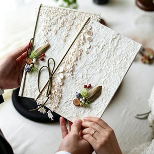 May include: A rectangular off-white textured book or folder featuring intricate cream-colored lace floral appliques, a dark green cord tied in a bow, and decorative accents consisting of small red berries, green pine needles, and brown wooden discs.