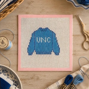 Könnte beinhalten: Ein Gobelin mit einem blauen Pullover-Design und den weißen Buchstaben "UNC". Die Leinwand ist rosa umrandet und liegt auf einer Holzoberfläche, umgeben von Nähutensilien wie Faden und Schere.