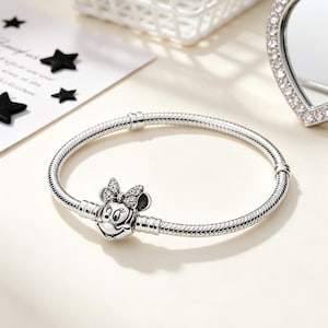 Puede incluir: Una pulsera plateada con un charm de Minnie Mouse. El charm presenta la cara de Minnie con un lazo adornado con peque&ntilde;as piedras transparentes. La pulsera tiene un dise&ntilde;o de cadena de serpiente y se muestra sobre una superficie clara.