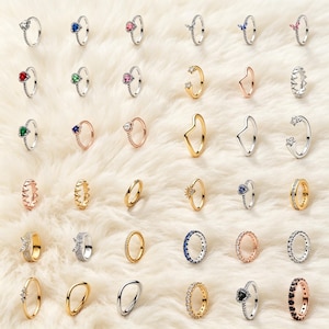 Può includere: Una collezione di vari anelli in metallo disposti in una griglia, caratterizzati da design con pietre preziose a forma di cuore, rotonde e a stella, incastonature pavé, fasce lisce e finiture testurizzate in tonalità argento, oro e oro rosa.