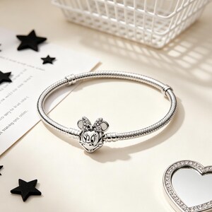 Puede incluir: Una pulsera plateada con un charm de Minnie Mouse. El charm presenta la cara de Minnie con un lazo y est&aacute; adornado con peque&ntilde;os acentos brillantes. La pulsera est&aacute; sobre una superficie clara con decoraciones en forma de estrella negra.