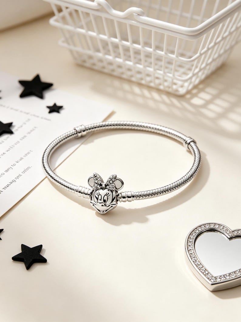 Puede incluir: Una pulsera plateada con un charm de Minnie Mouse. El charm presenta la cara de Minnie con un lazo y est&aacute; adornado con peque&ntilde;os acentos brillantes. La pulsera est&aacute; sobre una superficie clara con decoraciones en forma de estrella negra.