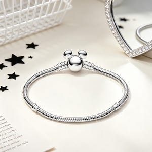 Puede incluir: Una pulsera plateada con un dije en forma de Mickey Mouse. La pulsera tiene un dise&ntilde;o de cadena de serpiente y se muestra sobre una superficie blanca. Un espejo en forma de coraz&oacute;n con borde de pedrer&iacute;a y una cesta blanca est&aacute;n en el fondo.