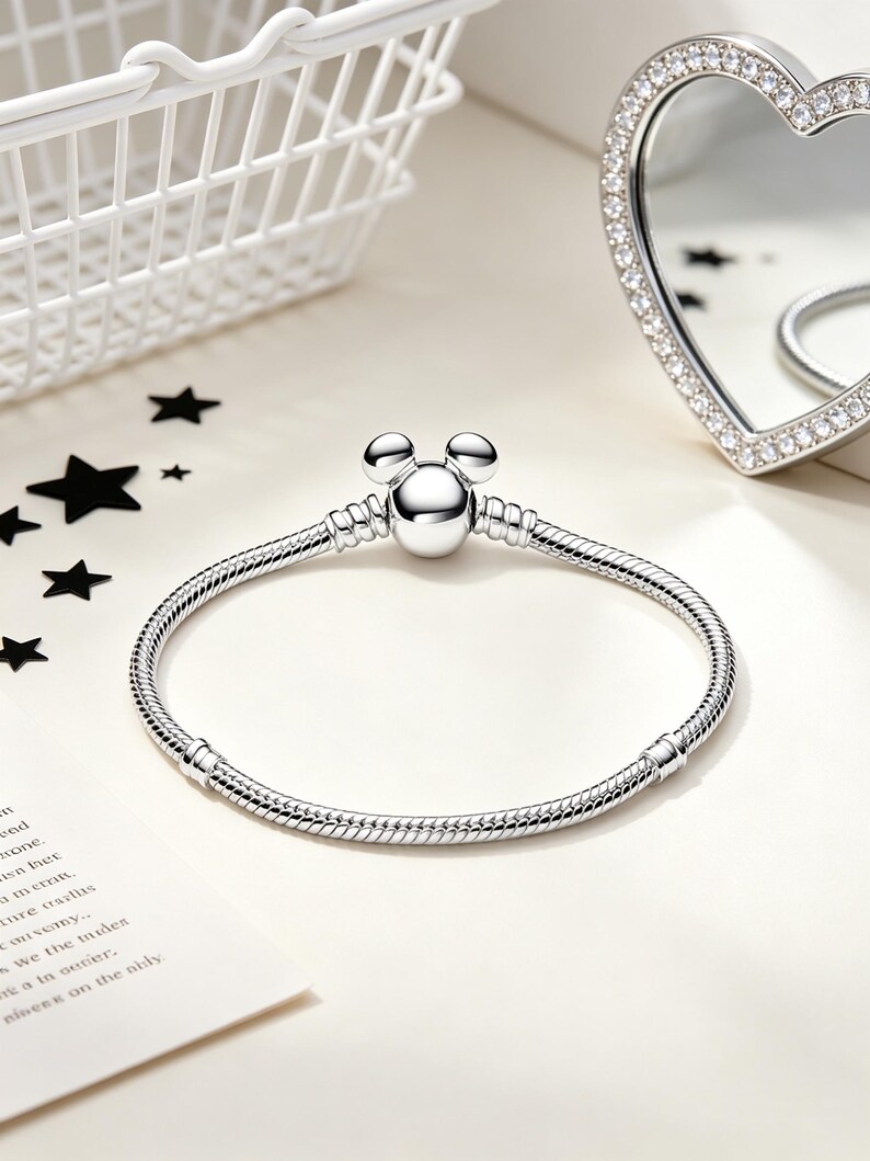 Puede incluir: Una pulsera plateada con un dije en forma de Mickey Mouse. La pulsera tiene un dise&ntilde;o de cadena de serpiente y se muestra sobre una superficie blanca. Un espejo en forma de coraz&oacute;n con borde de pedrer&iacute;a y una cesta blanca est&aacute;n en el fondo.