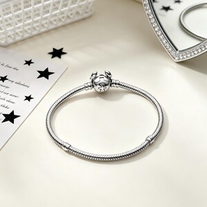 Puede incluir: Una pulsera plateada con un cierre en forma de cangrejo. La pulsera tiene un dise&ntilde;o de cadena de serpiente y se muestra sobre una superficie blanca. Un espejo en forma de coraz&oacute;n con un borde de diamantes est&aacute; en el fondo.