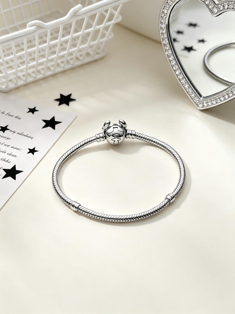 Puede incluir: Una pulsera plateada con un cierre en forma de cangrejo. La pulsera tiene un dise&ntilde;o de cadena de serpiente y se muestra sobre una superficie blanca. Un espejo en forma de coraz&oacute;n con un borde de diamantes est&aacute; en el fondo.