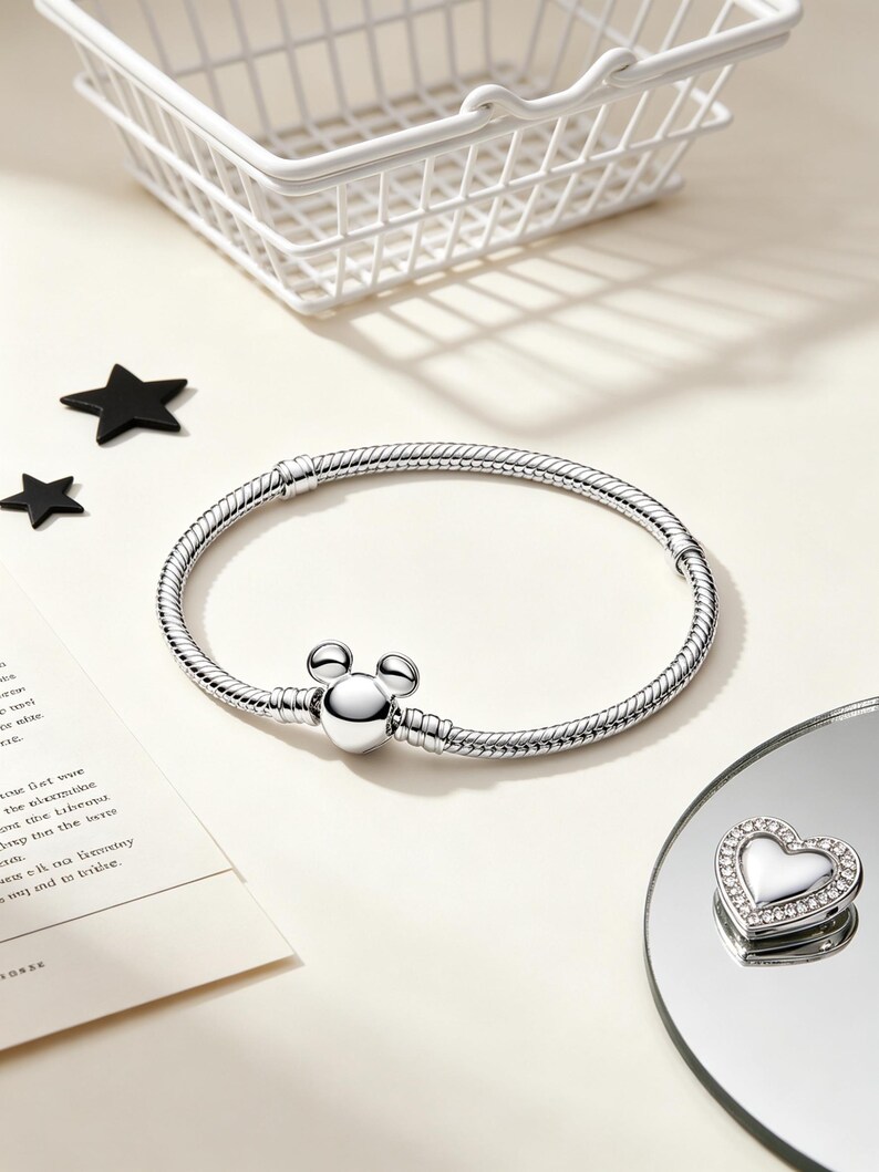Puede incluir: Una pulsera plateada con un cierre en forma de Mickey Mouse y un charm en forma de coraz&oacute;n con borde de cristal. La pulsera est&aacute; sobre una superficie blanca con una cesta de alambre blanca y decoraciones en forma de estrella negra.