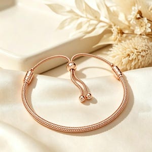 Puede incluir: Una pulsera de cadena de serpiente en tono oro rosa con un mecanismo de cierre deslizante ajustable. La pulsera tiene dos cadenas finas que se extienden desde una cuenta central, cada una terminada con una pequeña tapa redonda y flexible.