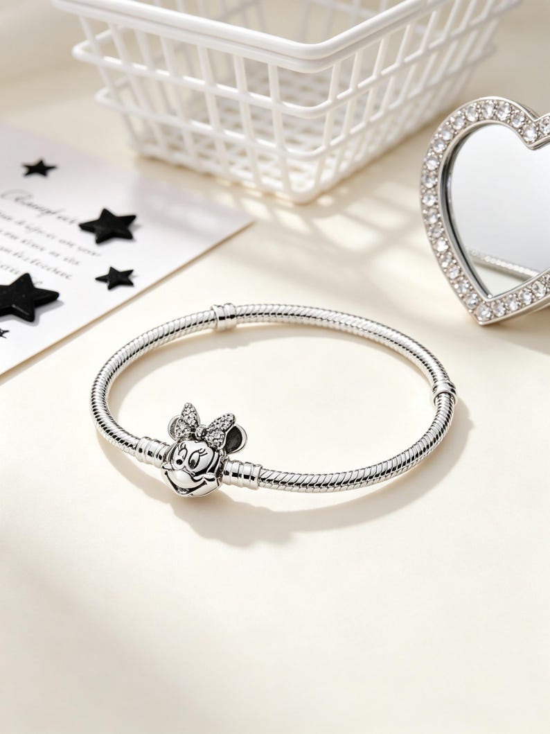 Puede incluir: Una pulsera plateada con un charm de Minnie Mouse. El charm presenta la cara de Minnie con un lazo adornado con peque&ntilde;as piedras transparentes. La pulsera tiene un dise&ntilde;o de cadena de serpiente y se muestra sobre una superficie clara.