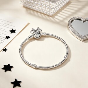 Puede incluir: Una pulsera plateada con un charm de Minnie Mouse con cara en forma de coraz&oacute;n y lazo. La pulsera tiene un dise&ntilde;o de cadena de serpiente. Una cesta blanca, un espejo en forma de coraz&oacute;n y confeti negro en forma de estrella tambi&eacute;n est&aacute;n en la imagen.