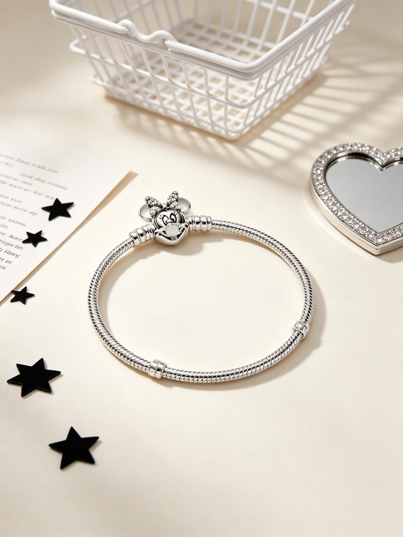 Puede incluir: Una pulsera plateada con un charm de Minnie Mouse con cara en forma de coraz&oacute;n y lazo. La pulsera tiene un dise&ntilde;o de cadena de serpiente. Una cesta blanca, un espejo en forma de coraz&oacute;n y confeti negro en forma de estrella tambi&eacute;n est&aacute;n en la imagen.