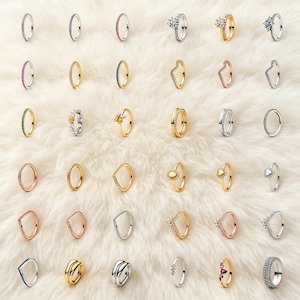 Puede incluir: Una colección de treinta anillos dispuestos en una cuadrícula, con diversos diseños que incluyen bandas pavé, engastes en forma de corazón, estilos de tiara en V y anillos con motivos florales, solares y de abejas en tonos plata, oro y oro rosa con gemas.