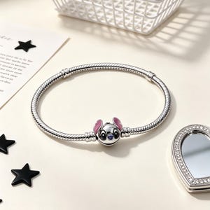 Puede incluir: Una pulsera plateada con un charm que representa un personaje de dibujos animados con orejas rosas y cara azul. La pulsera est&aacute; sobre una superficie clara, con un peque&ntilde;o espejo y decoraciones de estrellas negras.