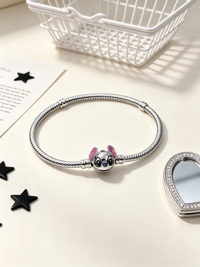 Puede incluir: Una pulsera plateada con un charm que representa un personaje de dibujos animados con orejas rosas y cara azul. La pulsera est&aacute; sobre una superficie clara, con un peque&ntilde;o espejo y decoraciones de estrellas negras.