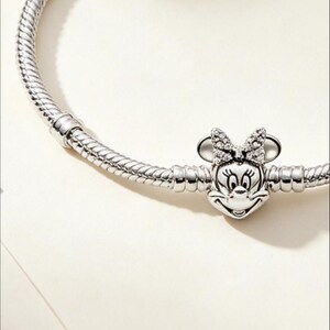 Puede incluir: Una pulsera de plata con un charm de Minnie Mouse. El charm presenta la cara y el lazo de Minnie, adornados con peque&ntilde;as piedras brillantes. La pulsera tiene un dise&ntilde;o de cadena de serpiente texturizada.