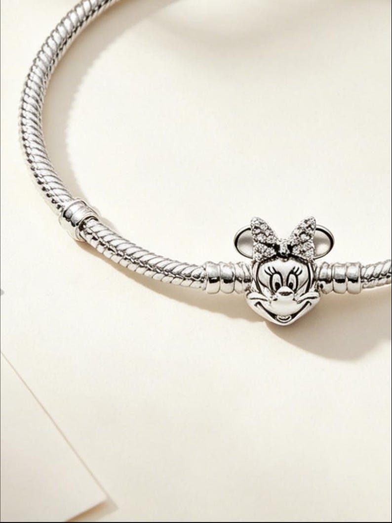 Puede incluir: Una pulsera de plata con un charm de Minnie Mouse. El charm presenta la cara y el lazo de Minnie, adornados con peque&ntilde;as piedras brillantes. La pulsera tiene un dise&ntilde;o de cadena de serpiente texturizada.
