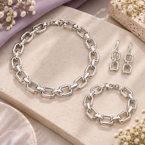 Può includere: Un set di gioielli color argento composto da una collana a maglie grosse, un bracciale a maglie abbinato e un paio di orecchini pendenti. Ogni pezzo presenta maglie rettangolari intrecciate e lucide con una finitura brillante.