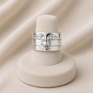 May include: A polished silver ring featuring a sculptural design of a human face. The band is wide and smooth, with embossed details depicting two eyes, a nose, and a mouth, creating a cohesive facial expression across the metallic surface.