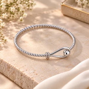 Può includere: Un bracciale rigido color argento caratterizzato da una fascia intrecciata a corda. Il bracciale presenta un meccanismo di chiusura a gancio che si fissa con una perla sferica liscia e lucida all'estremità dell'asola, creando un cerchio.
