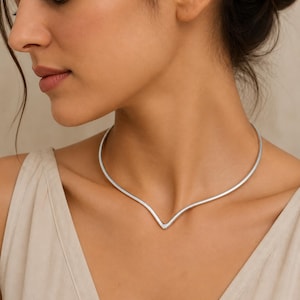 May include: A silver-toned metal choker necklace featuring a smooth, rigid band that curves into a distinct V-shape at the center. The necklace has a polished, reflective finish and sits closely around the base of the neck with a minimalist design.