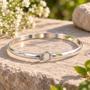 May include: A polished silver bangle bracelet featuring a smooth, rounded band that terminates in a central circular ring clasp. The metal surface reflects light, highlighting the clean, continuous lines of the circular jewellery piece.
