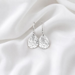 May include: A pair of silver drop earrings featuring a teardrop shape with a hammered, textured finish. Each earring hangs from a simple silver hook, reflecting light across the uneven, metallic surface of the teardrop pendant design.