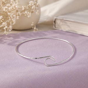 Puede incluir: Una pulsera rígida de plata que presenta una banda metálica fina y continua que se curva en forma de onda estilizada en la abertura. La superficie del metal plateado tiene una textura martillada sutil, creando un acabado reflectante.