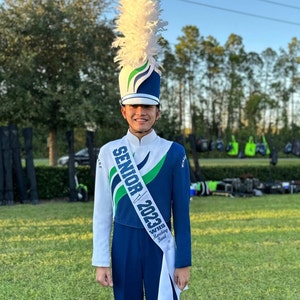 May include: A marching band uniform featuring a white and blue jacket with green accents, matching blue trousers, and a tall white feathered shako hat. A white sash across the chest displays the text SENIOR 2023 WHS Marching Band in blue.