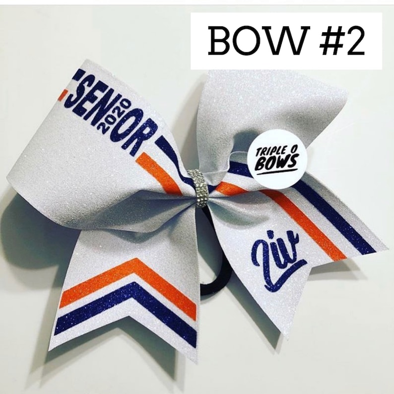 Puede incluir: Lazo de animadora blanco brillante con rayas naranjas y azul marino. El lazo tiene el texto "SENIOR 2020" y "Ziv". Un peque&ntilde;o logotipo redondo dice "TRIPLE O BOWS". El lazo est&aacute; atado con una banda de diamantes de imitaci&oacute;n.