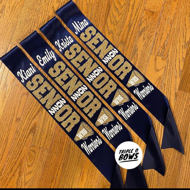 Puede incluir: Bandas de graduaci&oacute;n de sat&eacute;n azul marino con letras doradas con purpurina. Cada banda muestra "SENIOR" y el nombre de un estudiante en blanco. Las bandas tambi&eacute;n presentan "WHS Warriors" y un gr&aacute;fico de meg&aacute;fono. De Triple O Bows.