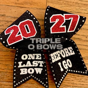 May include: A black glitter hair bow featuring the red glitter numbers 20 and 27. The text ONE LAST BOW BEFORE I GO is printed in white. The bow is embellished with small clear rhinestones along the edges and includes the text TRIPLE O BOWS.