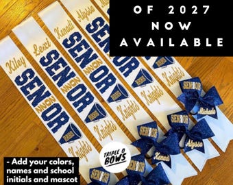 Puede incluir: Bandas de graduación blancas y azul marino con letras doradas y lazos de purpurina azul marino. Las bandas tienen la palabra "SENIOR" y los nombres de los graduados. También se ve el texto "CLASS OF 2027 NOW AVAILABLE".
