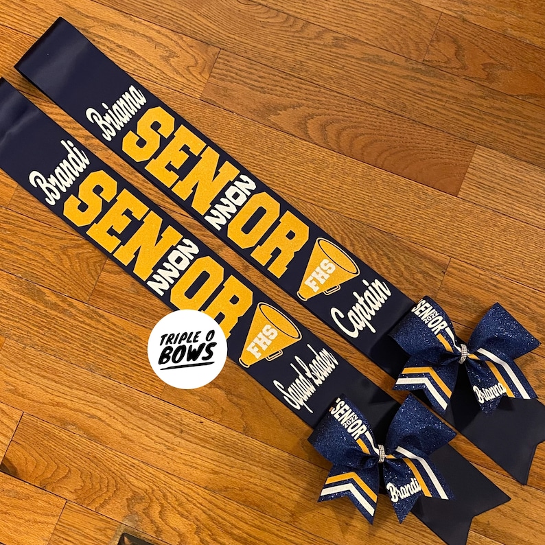 Puede incluir: Dos bandas de graduaci&oacute;n azul marino con letras doradas que dicen "SENIOR 2022". Cada banda tiene un lazo a juego con purpurina y los nombres "Brandi" y "Brianna". Una banda dice "Squad Leader" y la otra "Captain".