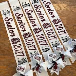 May include: Four white graduation sashes with maroon glitter text reading Senior 2027, individual names, and a megaphone graphic with JGHS. Each sash features a white hair bow with a graduation cap icon, maroon tassel, and the word Senior.