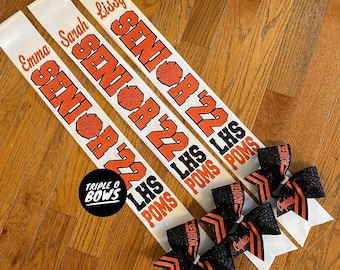 May include: Three white satin sashes with orange and black glitter text reading SENIOR '22 LHS POMS and individual names Emma, Sarah, and Lissy, accompanied by three black glitter hair bows with orange stripes and the word Captain printed on them.