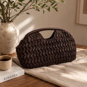 May include: A dark brown, woven handbag with a rounded shape and a top handle. The bag has a textured pattern and sits on a beige surface. A small cup of coffee and a book titled "KINFOLK" are in the foreground, with a vase of greenery in the background.