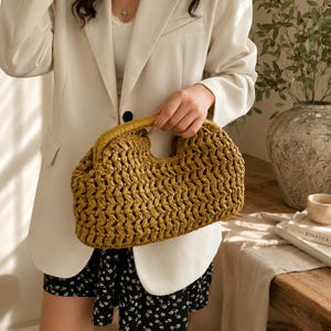 May include: A woven straw handbag with a curved handle. The bag is a golden-yellow color and has a textured, open weave design. The handle is a lighter shade of yellow. The bag is held by a person wearing a white blazer and black and white shorts.